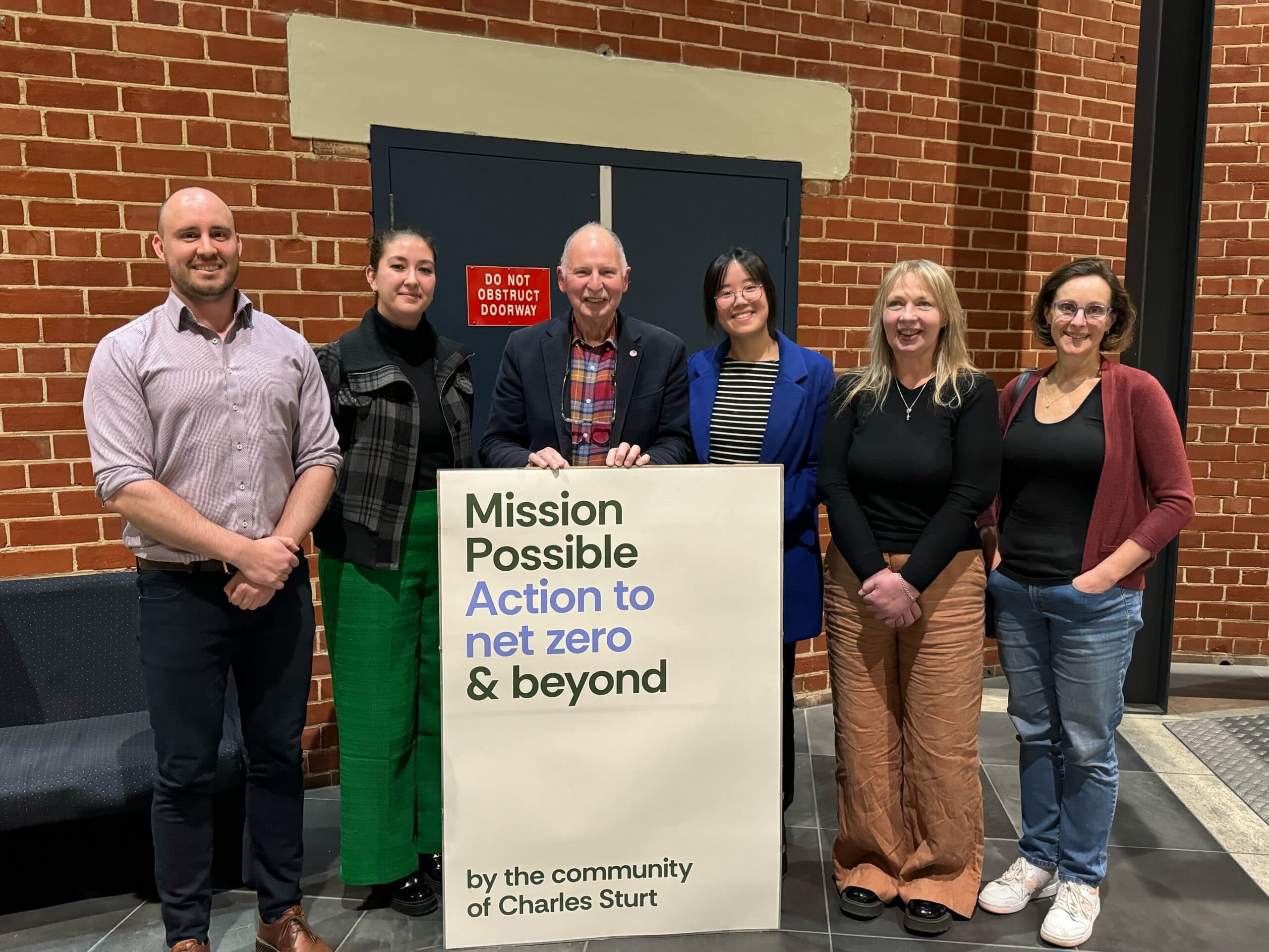 Six Mission Possible members standing together with a sign that reads 'Mission Possible — Action to net zero & beyond, by the community of Charles Sturt'.